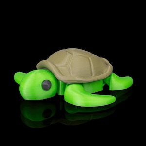 May include: A 3D-printed toy turtle with a light green body and a tan shell. The turtle has a detailed shell pattern and black eye details. The toy is set against a black background.