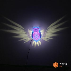Puede incluir: Una luz nocturna en forma de búho de color morado y azul proyecta una sombra de alas en la pared. La luz está hecha de formas geométricas y tiene un cable blanco.