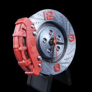 Peut inclure: Une horloge originale avec un disque de frein argenté et des chiffres rouges à 12, 3 et 6 heures. Un étrier de frein rouge est fixé sur le côté. L'horloge a des aiguilles noires et est montée sur un support noir.