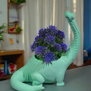 Op de afbeelding: Een lichtgroene dinosaurusvormige plantenpot met een paarse bloemschikking erin. De pot staat op een grijs oppervlak.