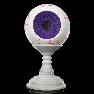Puede incluir: Un globo ocular blanco con venas rojas y un iris morado sobre un pedestal blanco. El globo ocular es una decoración de Halloween.