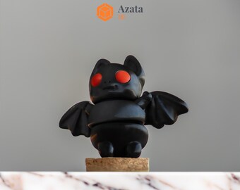 May include: A black bat figurine with red eyes, standing on a cork base. The figurine is made of plastic and has a segmented body. The bat has wings that are spread out to the sides.