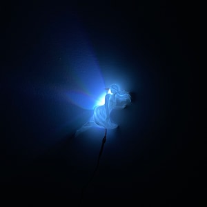 May include: A white fairy figurine with wings, illuminated by a blue light, against a dark background.