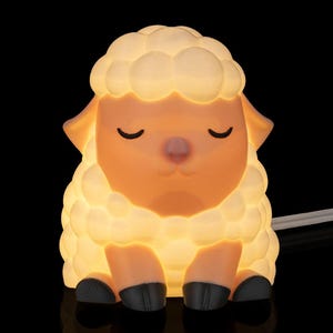 May include: A cute sheep-shaped night light, glowing with a soft, warm light. The lamb has a fluffy, white wool body, a peach-coloured face and ears, and black hooves. The eyes are closed, and the nose is pink. The night light is illuminated.
