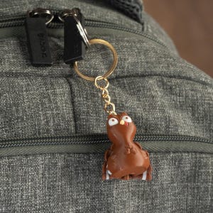 Puede incluir: Un llavero en forma de pavo marrón con detalles blancos y ojos saltones cuelga de una cadena y un anillo dorados unidos a una mochila gris.