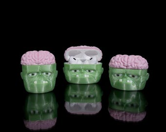 May include: Three green Frankenstein-themed AirPod cases with pink brain-shaped lids. One case is open, revealing white earbuds. The cases have a glossy finish and are set against a black background. The Azata 3D logo is in the bottom right corner.