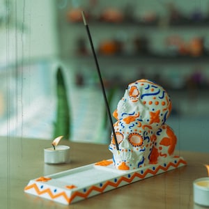 Puede incluir: Quemador de incienso de cerámica blanca con forma de tres calaveras de azúcar apiladas con diseños florales naranja y azul. El quemador tiene un patrón de zigzag blanco y naranja alrededor del borde.
