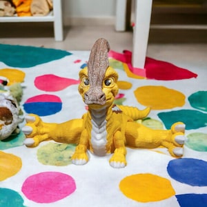 May include: A yellow dinosaur toy with brown and white markings sits on a white and colourful play mat. The dinosaur has a long neck and a long tail. It has large, round eyes and a friendly expression.