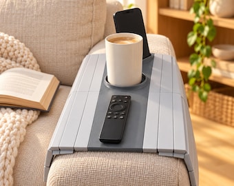 May include: A light gray flexible sofa armrest tray with a dark gray center panel. The tray features a circular cup holder containing a white mug, a slot holding a black smartphone, and a black remote control resting on the flat surface.