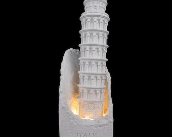 May include: A white stone-textured sculpture of the Leaning Tower of Pisa set against a jagged rock-like backdrop. The base features the word ITALY engraved in capital letters. A warm yellow light illuminates the structure from behind the tower.