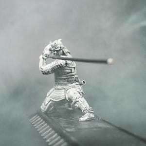 Puede incluir: Una figura de plata de un guerrero samurái en posición de combate, sosteniendo una espada. La figura está de pie sobre una base negra con una superficie texturizada.