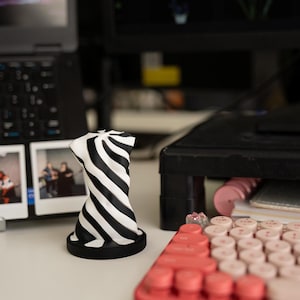 May include: A black and white striped 3D printed sculpture on a black base. The sculpture is twisted and has a swirling pattern. The sculpture is on a white surface with a red keyboard in the foreground.