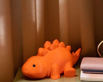 May include: Orange dinosaur figurine with a spiky back and large, round eyes. The figurine is sitting on a light green nightstand.
