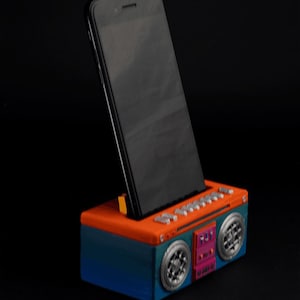 Può includere: Un supporto per telefono in stile boombox blu e arancione con uno smartphone nero nel supporto. Il supporto ha una griglia dell'altoparlante argentata e un pannello di controllo rosso e rosa.