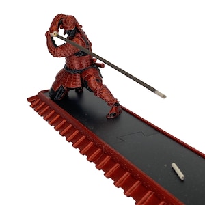 Può includere: Statuetta di samurai rossa e nera che tiene una spada, posizionata su una base rettangolare nera con un bordo rosso a forma di tetto. La statuetta è dettagliata con armatura ed elmo. Un bastoncino di incenso è nella mano del samurai.
