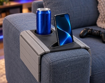 May include: A gray flexible sofa armrest tray with a dark gray center insert featuring a circular cup holder containing a blue aluminum beverage can and a triangular slot holding a black smartphone with a blue abstract screen display.