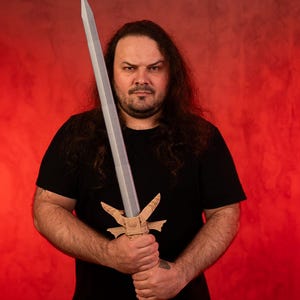 May include: A man with long brown hair holds a silver sword with a wooden handle and decorative details. He is wearing a black t-shirt and standing in front of a red background.