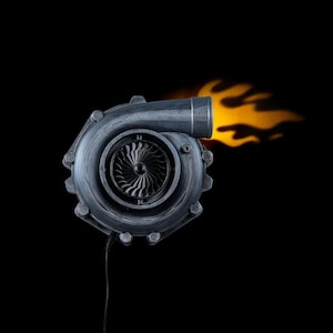 Op de afbeelding: Een grijze turbocharger met een vlameffect dat uit de uitlaat komt. De turbocharger heeft een gedetailleerd ontwerp met een centrale ventilator en boutdetails. Een zwart netsnoer loopt van de onderkant.