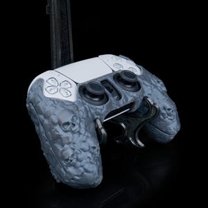 May include: A white PlayStation 5 DualSense controller with a gray skull-patterned grip. The controller is attached to a black stand.