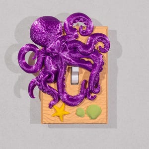Könnte beinhalten: Ein Lichtschalter-Abdeckung mit einem lila Oktopus auf einem sandbraunen Hintergrund. Der Oktopus hat acht Tentakel und befindet sich über einem Lichtschalter. Der Hintergrund zeigt einen gelben Seestern und zwei grüne Muscheln.