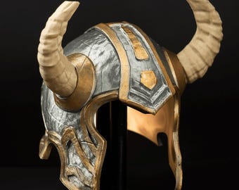 May include: A horned helmet featuring a metallic silver finish with gold-colored trim and accents. The helmet includes two large, curved, beige-colored horns extending from the sides and a central ridge running from the front to the back.