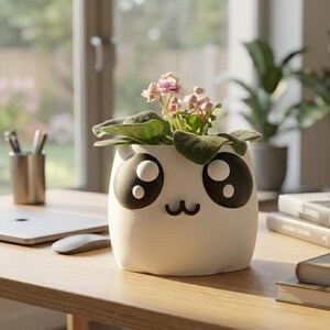 Pode incluir: Um vaso de cerâmica branco com um design de rosto de desenho animado, com olhos pretos grandes e uma boca preta pequena. O vaso contém uma planta florida com folhas verdes e pequenas flores rosa. O vaso está em uma mesa de madeira.