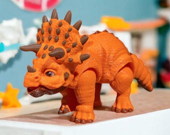 May include: Orange and brown toy dinosaur with a frill and horns. The dinosaur is standing on a white table.