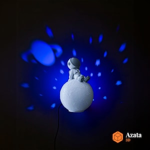 Op de afbeelding: Een wit astronautje zit op een wit maanvormig nachtlampje, dat blauwe sterren en een blauwe planeet projecteert op een donkere achtergrond. Het nachtlampje heeft een snoer.