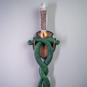 Può includere: Una spada verde a forma di serpente con un manico in legno e un occhio dorato sul pomo. La spada è montata su una placca di legno con un design decorativo.
