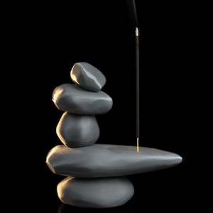 May include: A gray stone incense holder with a stack of smooth, rounded stones. An incense stick is lit, with smoke rising against a black background. The holder is designed to resemble a zen garden.
