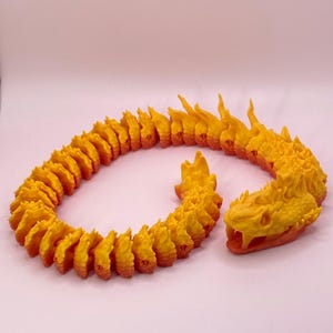 Puede incluir: Una serpiente impresa en 3D hecha de plástico naranja y amarillo. La serpiente está enroscada en un círculo con la cabeza sobresaliendo. La serpiente tiene una cabeza detallada con colmillos y escamas.