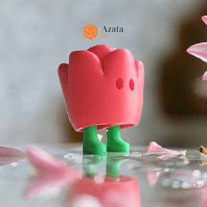 May include: A pink 3D printed rose with green legs standing on a reflective surface. The rose has two black eyes. The text "Azata 3D" is visible in the background.