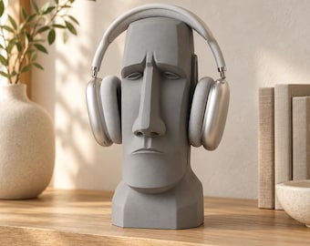 May include: A matte gray Moai statue head functioning as a headphone stand. The sculpture features a stylized face with a prominent nose, heavy brow, and closed mouth. A pair of silver over-ear wireless headphones rests on the head of the statue.