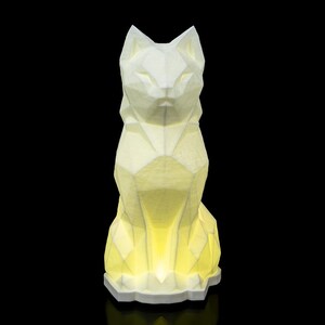 Peut inclure: Lampe en forme de chat blanc géométrique avec un intérieur jaune brillant. La lampe a un design à facettes, lui donnant un aspect moderne et artistique. Le chat est assis droit, avec des oreilles pointues et un regard concentré. La lampe repose sur une base blanche.