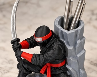 May include: A black and red ninja figurine pen holder featuring a crouching figure holding a silver curved sword. The figure is positioned in front of a gray, rock-textured cylindrical container that holds two metallic silver pens.