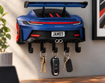 May include: A blue wall-mounted key holder shaped like the rear of a Porsche GT3RS sports car. It features a large rear wing with the text PORSCHE, a license plate reading JAMES, dual exhaust pipes, and five black hooks holding various keys.