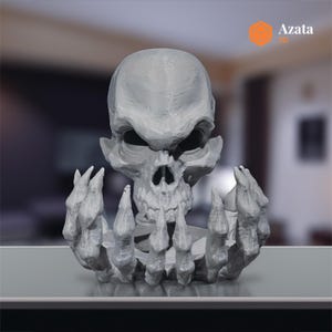 Puede incluir: Un cráneo gris impreso en 3D con una mano esquelética sosteniéndolo. El cráneo tiene una expresión amenazante con ojos hundidos y una boca ancha.