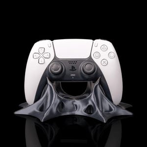 May include: A white and black PlayStation 5 controller resting on a dark gray, stylized stand. The controller features a symmetrical design with various button symbols and a PlayStation logo. The stand has an intricate, organic shape.