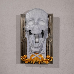 May include: A white skull light switch cover with a gold frame and crossbones design. The skull is open-mouthed and has a light switch in the mouth.