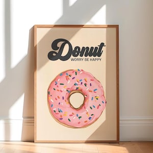 Könnte beinhalten: Ein gerahmter Druck mit einem rosa Donut mit bunten Streuseln. Das Wort "Donut" steht über dem Donut, darunter "Worry be happy". Der Druck hat einen hellbeigen Hintergrund und einen Holzrahmen. Das Bild ist vor einer weißen Wand.