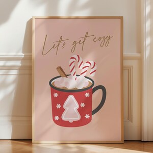 Könnte beinhalten: Ein rosa Poster mit dem Text "Let's get cozy" in goldenen Buchstaben. Eine rote Tasse mit weißen Schneeflocken und einem kleinen rosa Weihnachtsbaum ist gefüllt mit heißer Schokolade, die mit Marshmallows und zwei Zuckerstangen belegt ist.