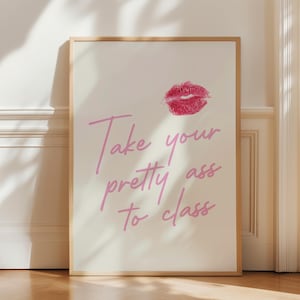 May include: A framed print with a light background features a pink lip print and pink cursive text that reads, "Take your pretty ass to class." The frame is a light wood color, and the print is displayed on a wooden floor.