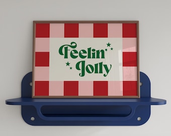 May include: A rectangular framed print featuring a red and light pink checkered border surrounding a white center. The text "Feelin' Jolly" is printed in dark green cursive lettering, accompanied by small dark green star icons on the white background.