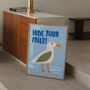 May include: A blue and white poster with a cartoon seagull holding a french fry in its beak. The poster has the text "Hide Your Fries!" in blue letters.