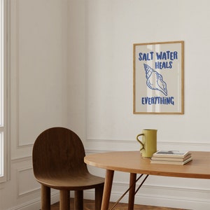 May include: A framed print with a seashell and the text "Salt Water Heals Everything" in blue lettering on a striped background. The print is hanging on a white wall above a wooden table with a mug and books.