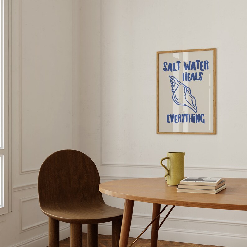 May include: A framed print with a seashell and the text "Salt Water Heals Everything" in blue lettering on a striped background. The print is hanging on a white wall above a wooden table with a mug and books.