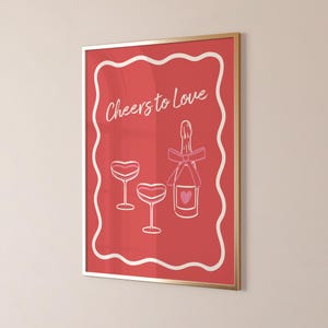 K&ouml;nnte beinhalten: Ein roter gerahmter Druck mit einem wei&szlig;en Rahmen und dem Text "Cheers to Love" in wei&szlig;er Schrift. Der Druck zeigt eine rosafarbene Champagnerflasche mit einer rosafarbenen Schleife und zwei rosafarbene Champagnergl&auml;ser.