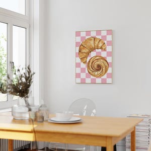 K&ouml;nnte beinhalten: Ein Aquarellbild eines Croissants und eines Zimtschnecken auf einem rosa-wei&szlig; karierten Hintergrund.