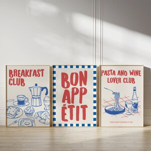 Op de afbeelding: Drie ingelijste kunstprints met rode en blauwe illustraties op een cr&egrave;mekleurige achtergrond. De prints luiden "BREAKFAST CLUB", "BON APP&Eacute;TIT" en "PASTA AND WINE LOVER CLUB". De pastaprint bevat de tekst "FRIDAY NIGHT STARTING AT 7PM".