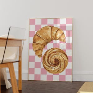 K&ouml;nnte beinhalten: Eine Aquarell-Illustration eines Croissants und eines Strudelgeb&auml;cks auf einem rosa-wei&szlig; karierten Hintergrund.